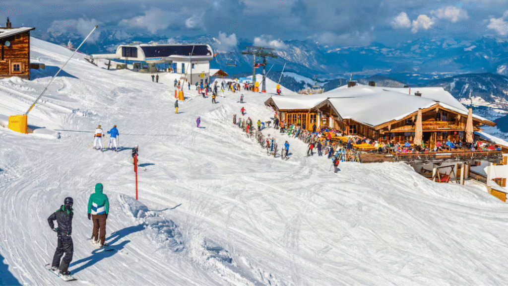 Skiers gather outside a mountainside restaurant at Alpbach ski resort, with gentle snowy slopes and ski lifts in the background.