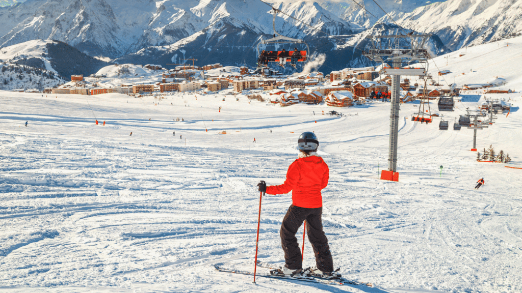 A skier stands at the top of a gentle slope overlooking the Alpe d’Huez ski resort, with cable cars, snow-covered runs, and mountain peaks in the background.