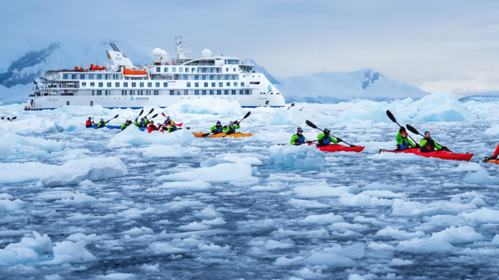 Aurora Expeditions cruise ship in icy Antarctic waters, with passengers kayaking among floating ice and snow under a cloudy sky.