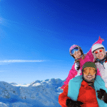 A happy family poses together on a sunny mountain with snowy peaks in the background, illustrating the Best European ski resorts for beginners.