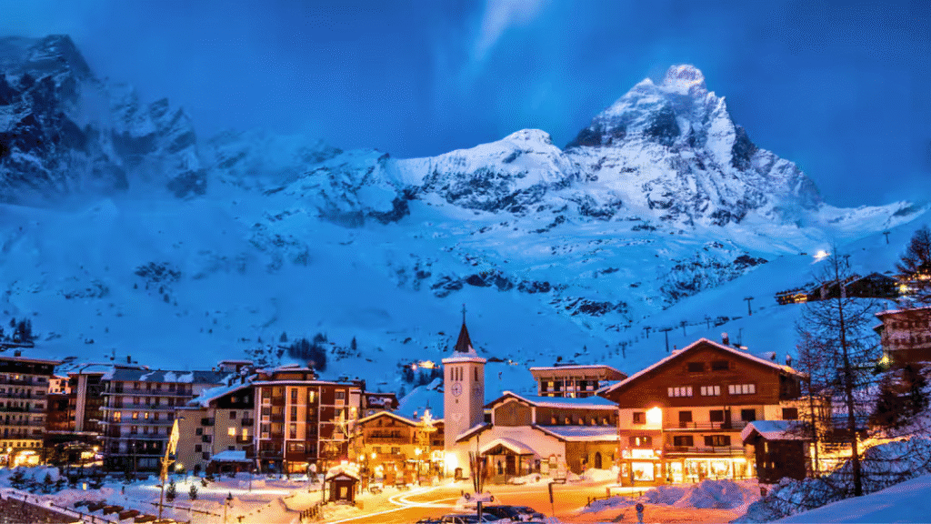 Cervinia ski resort at dusk, with warmly lit alpine buildings in the foreground and the snow-covered Matterhorn towering behind.