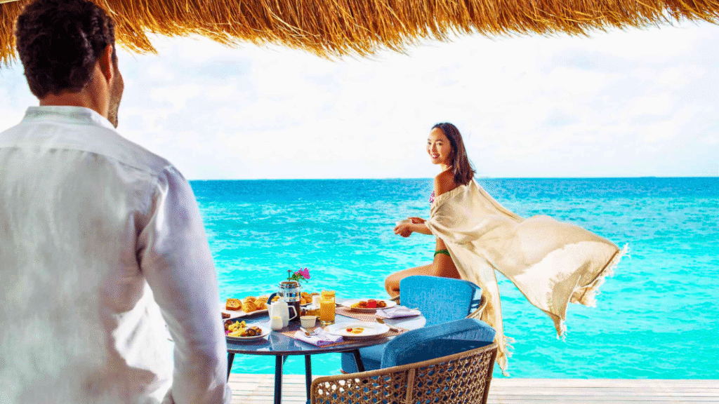A couple enjoys breakfast by the turquoise sea at an overwater villa at Emerald Maldives Resort and Spa, with tropical food and a stunning ocean view.