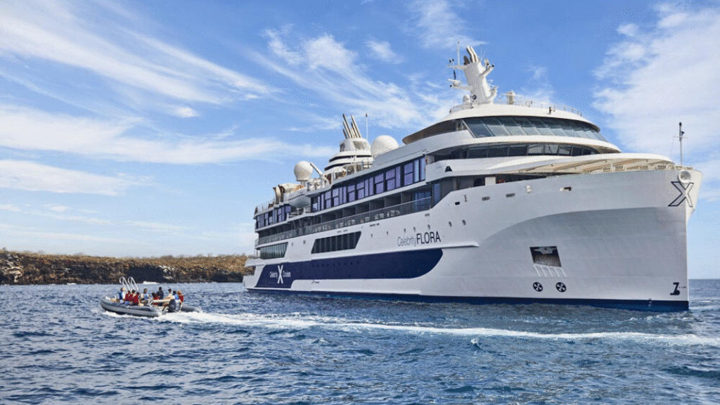 A modern expedition cruise ship, the Celebrity Flora, anchored near the coast with a small group of passengers boarding a zodiac boat in clear blue water under a partly cloudy sky.