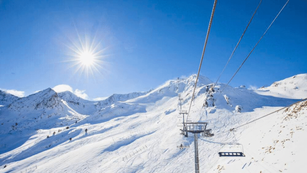 A ski lift rises over the sunlit, snow-covered mountains of Grandvalira ski resort with clear blue skies overhead.
