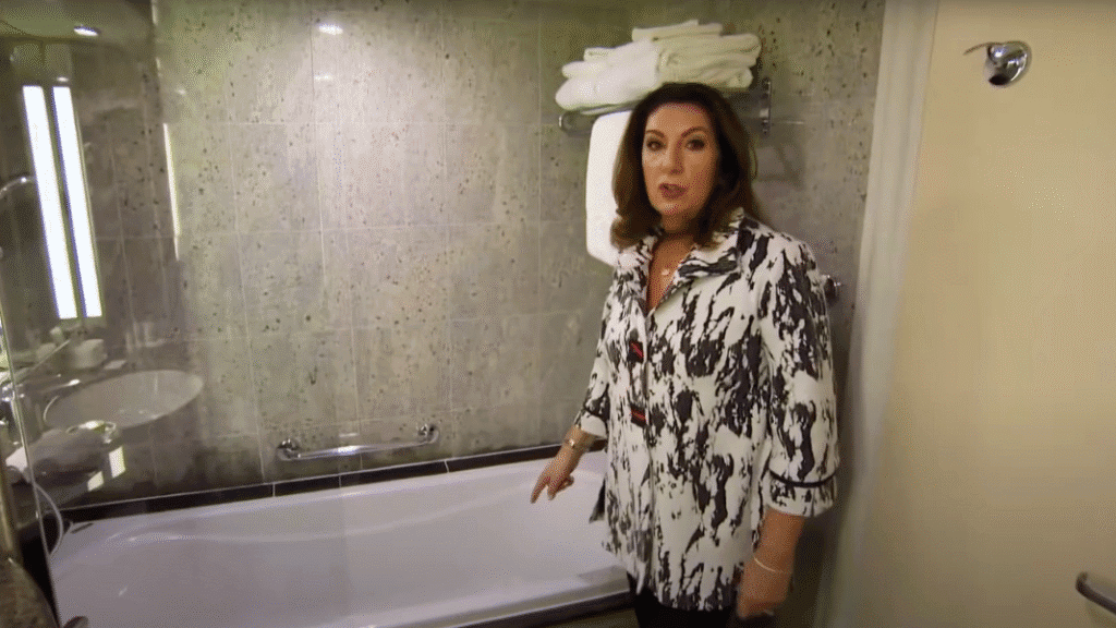 Jane McDonald stands beside a bathtub in a river cruise cabin bathroom in Europe, pointing as she offers practical advice.