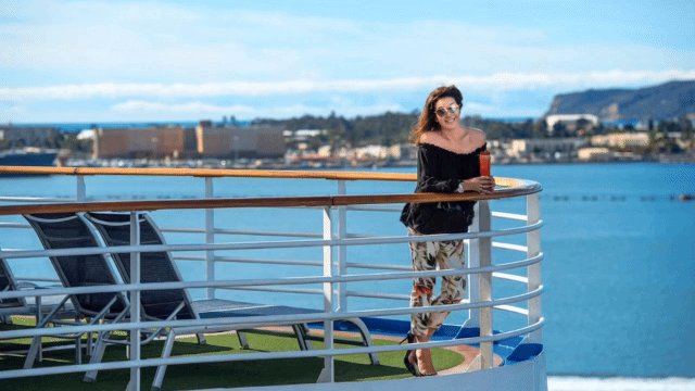 Jane McDonald stands smiling on the sun deck of a river cruise ship, enjoying a drink with scenic views in the background.