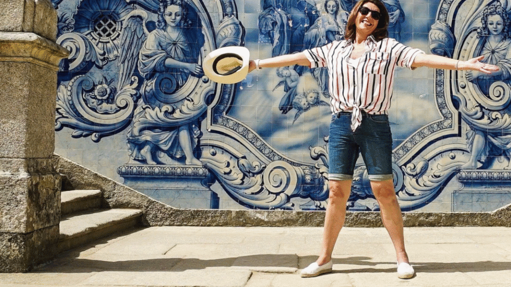 Jane McDonald stands with arms outstretched and smiling in front of a blue and white tiled mural, capturing the joy of her European river cruise journey.