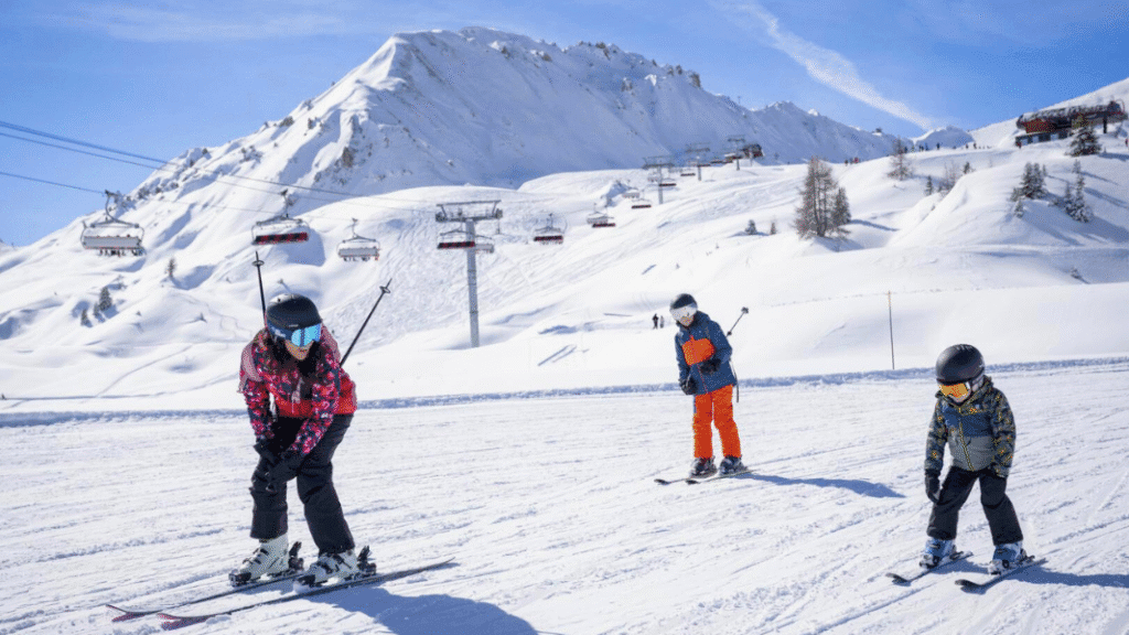 Three children learning to ski on a gentle, snow-covered slope at La Plagne ski resort, with chairlifts and safe play zones in the background.