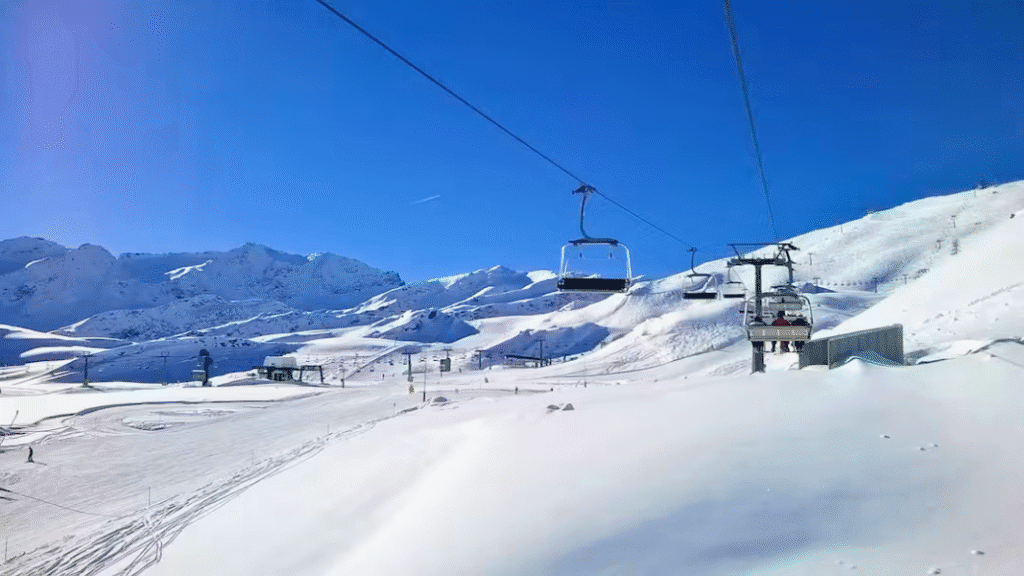 A chairlift ascends above the snow-covered slopes of La Thuile ski resort, set against clear blue skies and mountain peaks.