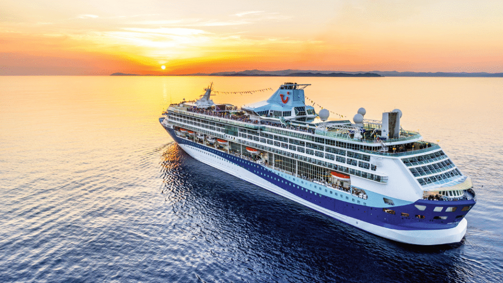 A Marella Cruises ship sails on calm waters at sunset, with the sun low on the horizon and a gentle orange glow reflecting off the sea.
