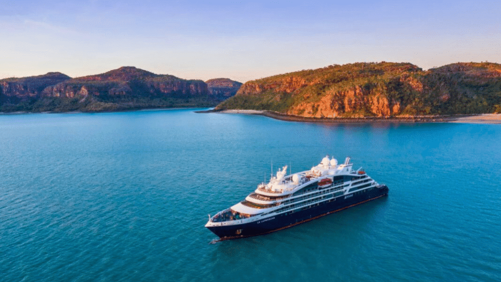 A luxury expedition cruise ship anchored in tranquil turquoise waters near a rugged, scenic coastline with rocky hills and sparse vegetation, under a clear sky.