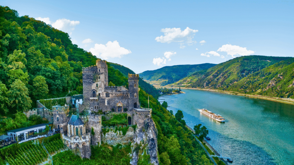 Medieval castle perched above the scenic Rhine River with a river cruise ship sailing past lush green hills, illustrating the storybook landscapes of European river cruising.
