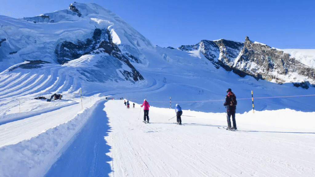 Skiers descend a wide, gentle piste at Saas-Fee ski resort, surrounded by high snowy mountains and glaciers under a clear blue sky.