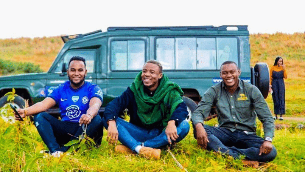 Three cheerful safari guides sit on the grass in front of a safari vehicle, with a woman walking in the background.
