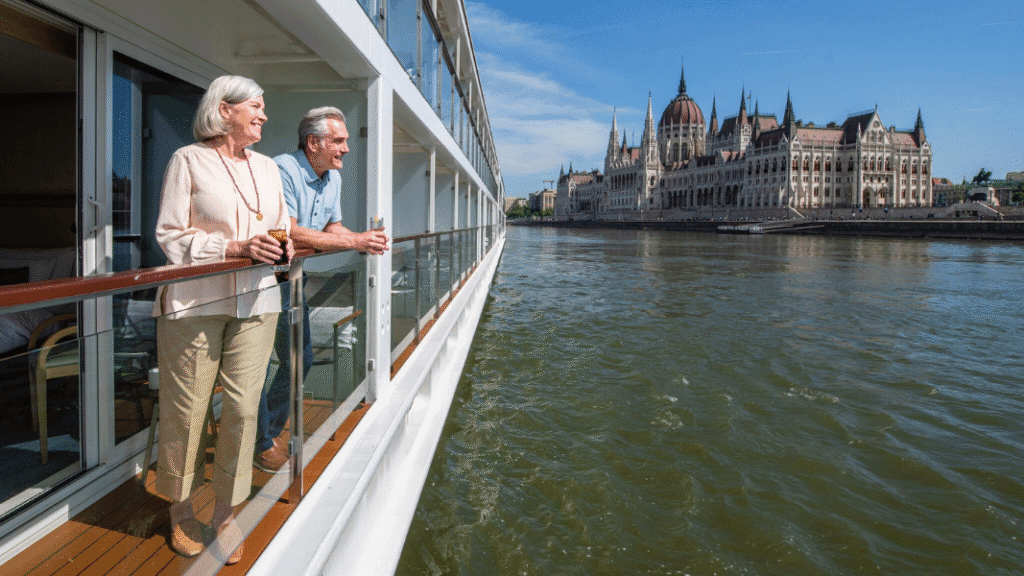 An older couple stands on the balcony of a river cruise ship, enjoying views of the Hungarian Parliament Building along the Danube River.